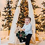 Jadiel is registered to the contest to win money with this photo: black_pants, carpet, child, christmas_tree, cute, decorations, festive, greenery, happy, holiday_decor, indoor, lights, pinecones, portrait, ribbon, rocking_horse, smiling, toddler, white_shirt, wooden_toy