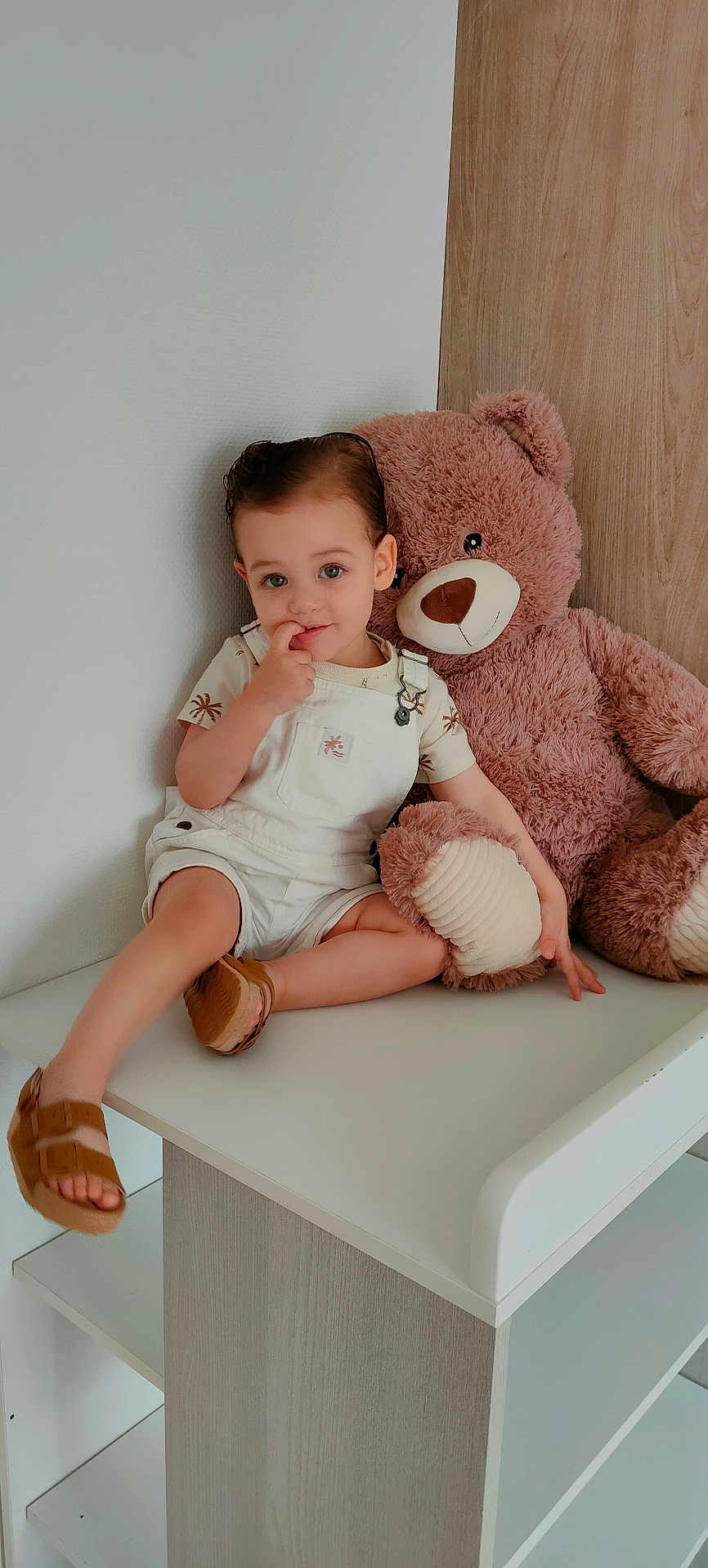 Anes participe au concours pour gagner de l'argent avec cette photo : toddler, child, plush_toy, teddy_bear, white_overalls, brown_sandals, finger_in_mouth, curious_expression, indoor, changing_table, minimalist_room, wood_panel, white_wall, sitting, cute, soft_lighting, comfortable, young_child, person, portrait