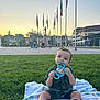 baby, child, grass, blanket, toy, outdoor, sunset, park, flags, building, curious, sitting, playful, greenery, person, footwear, casual_clothing, public_space, daylight, relaxation