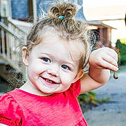Aaliyah is registered to the contest to win money with this photo: child, toddler, smile, happy, blue_eyes, red_shirt, hair_bun, outdoor, daylight, holding, small_object, face, person, cute, joyful, skin, expression, background, nature, casual_clothing