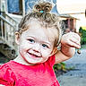 child, toddler, smile, happy, blue_eyes, red_shirt, hair_bun, outdoor, daylight, holding, small_object, face, person, cute, joyful, skin, expression, background, nature, casual_clothing