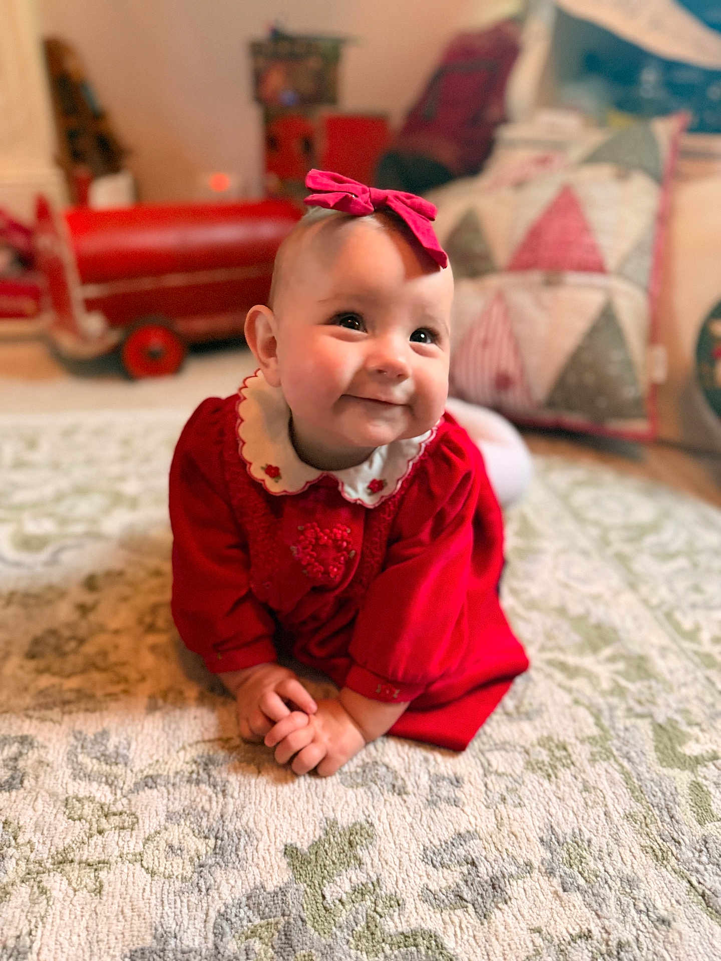 Hattie-Bloom joined the competition — help win amazing prizes! baby, infant, smiling, red_dress, bow, carpet, crawling, indoors, toy_car, pillow, patterned_rug, cute, child, happy, portrait, home, decor, person, warm_lighting, cozy
