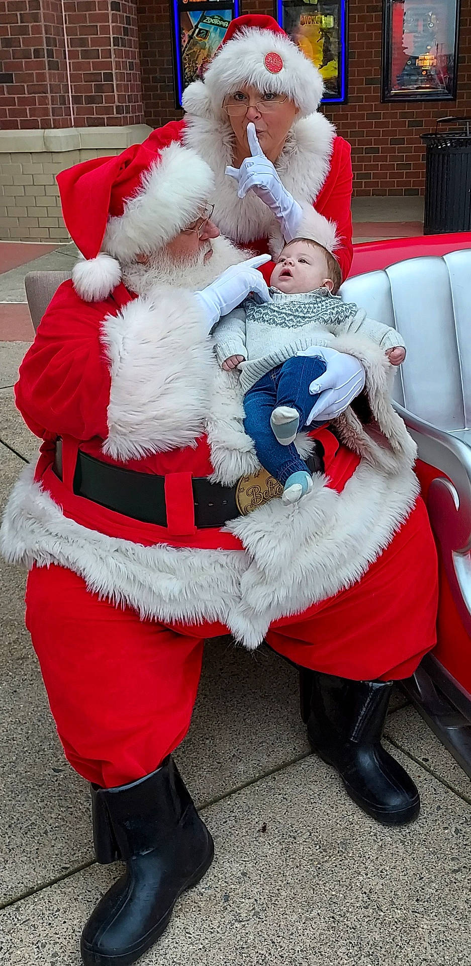 William is registered to the contest to win money with this photo: baby, bench, boots, brick_wall, child, christmas, costume, festive, glasses, gloves, hat, holiday, movie_posters, mrs_claus, outdoor, person, red_clothing, santa_claus, smile, white_fur