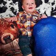 William joined the competition — help win amazing prizes! baby, smiling, plaid_shirt, denim_pants, barefoot, cushion, plush, cow_print, blue_cushion, indoor, portrait, happy, child, person, seated, cozy, fabric, cute, infant, colorful