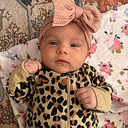 Emersyn is registered to the contest to win money with this photo: baby, infant, headband, leopard_print, onesie, pink_bow, blanket, floral_pattern, rug, fist, face, lying_down, indoor, child, cute, expression, clothing, portrait, young, person