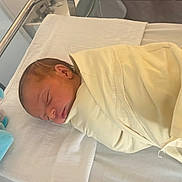 éline participe au concours pour gagner de l'argent avec cette photo : newborn, baby, swaddled, blanket, sleeping, hospital, bassinet, plush_toy, blue, infant, child, peaceful, resting, soft, cute, healthcare, bed, person, nursery, quiet