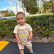 Kayden is registered to the contest to win money with this photo: child, toddler, baby, standing, striped_overalls, lion_patch, white_tshirt, blue_shoes, sneakers, hedge, bushes, trees, palm_tree, sky, clouds, asphalt, parking_lot, sidewalk, smile, outdoor