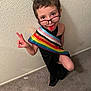 child, glasses, tongue, peace_sign, pride_flag, short_hair, black_cloth, carpet, wall, playful, indoor, person, portrait, happy, expression, standing, colorful, cute, fun, eyes