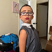 Glenna is registered to the contest to win money with this photo: child, glasses, makeup, lipstick, eyebrows, sweater_vest, tie, indoor, room, laundry_basket, clothes, door, cardboard_boxes, carpet, wall_art, person, portrait, fashion, expression, posing