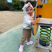 Amelia a rejoint le concours — aidez-le/la à gagner de superbes lots ! bench, building, child, cloudy_sky, curious, green_spring, hoodie, outdoor, pants, person, pink_bow, play, playground, sand, shoes, spring_ride, standing, toddler, trees, yellow