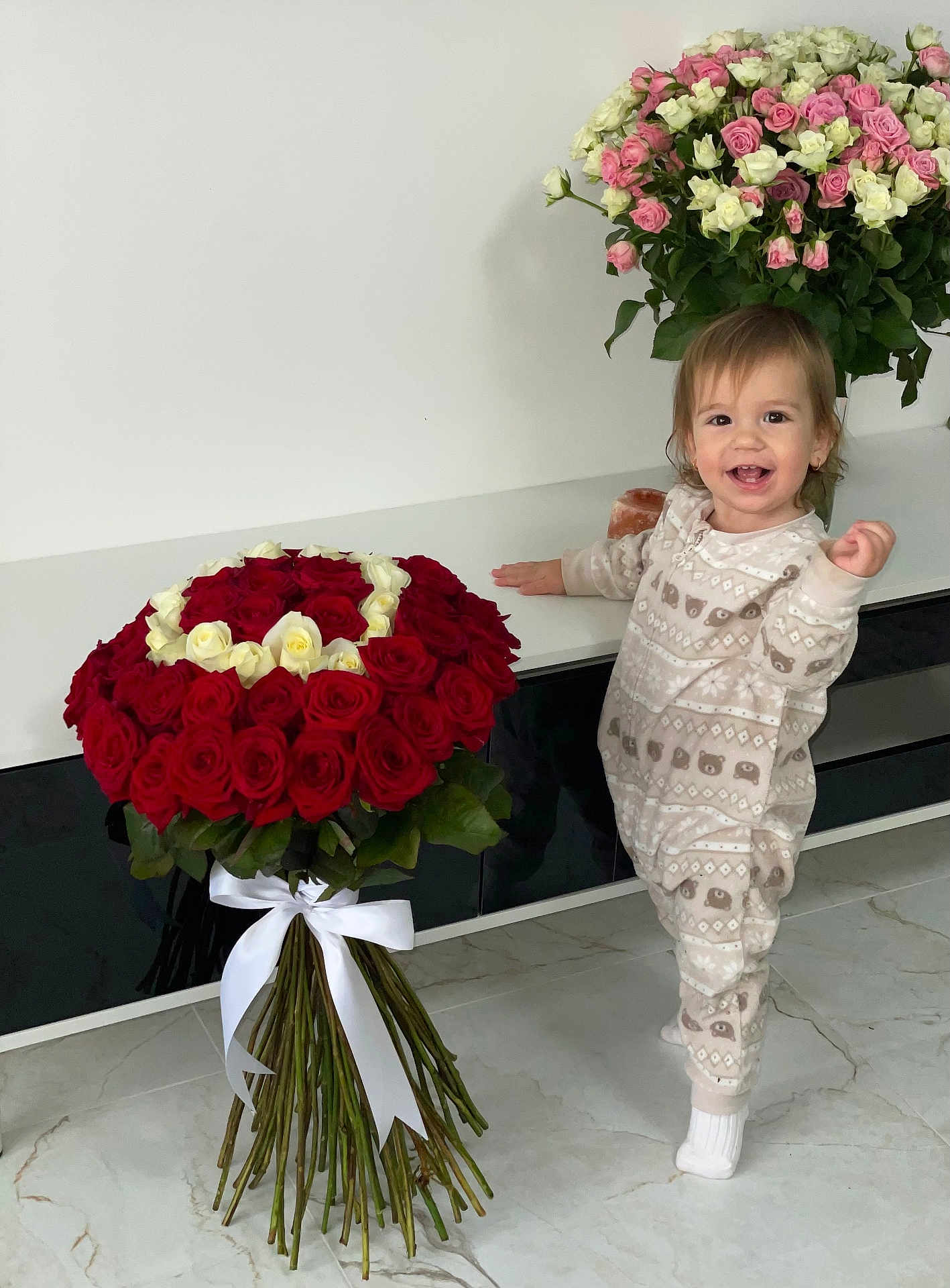 Amelia a rejoint le concours — aidez-le/la à gagner de superbes lots ! bouquet, cabinet, child, cute, decor, floor, flowers, green_leaves, happy, heart_shape, indoor, marble_floor, pajamas, person, plant, roses, smiling, standing, toddler, white_ribbon