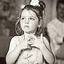 child, girl, portrait, black_and_white, dress, hair_bow, hands, thoughtful_expression, eyes, candid, indoor, blurred_background, adult, spectator, family, ceremony, soft_lighting, closeup, floral_pattern, innocence