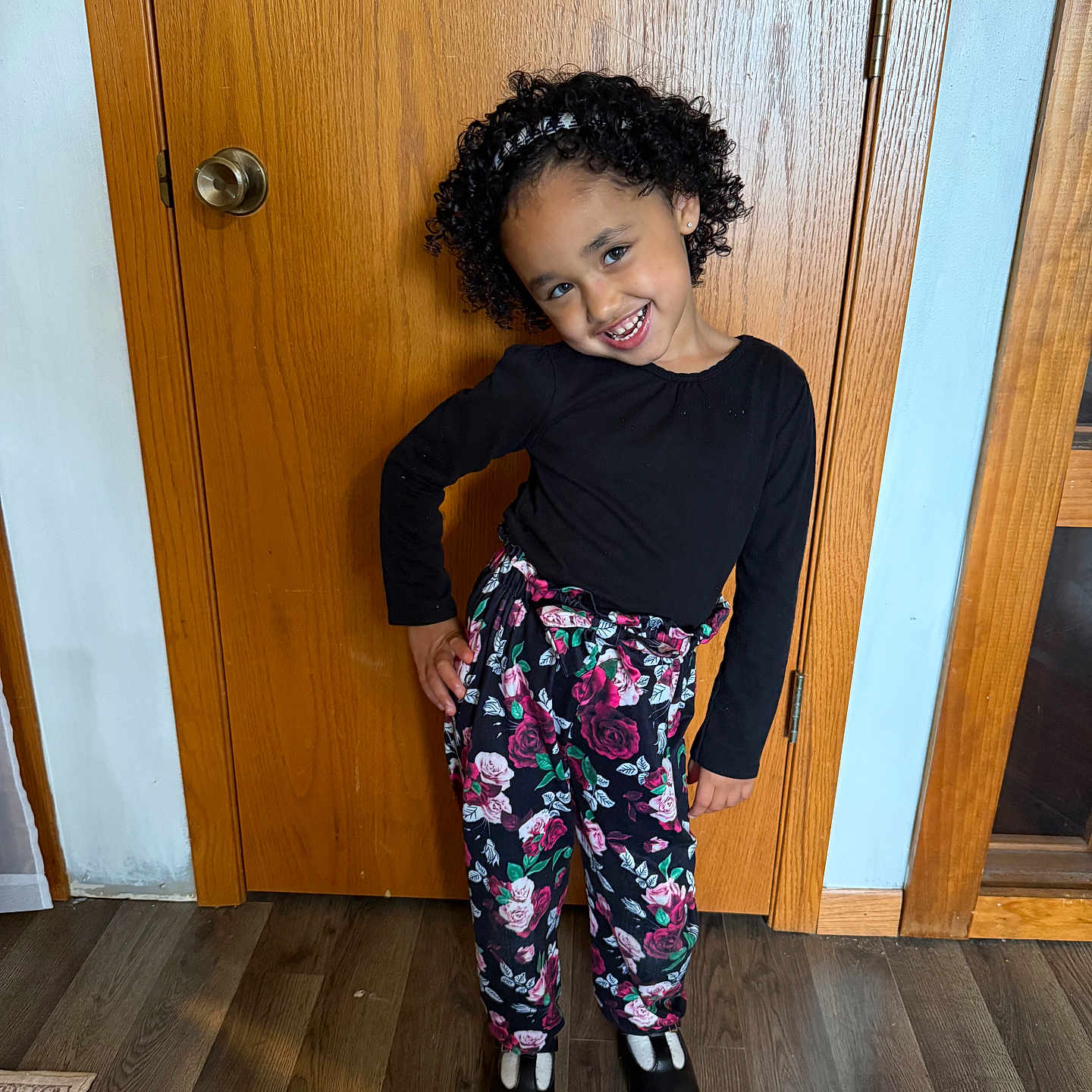 Serenity joined the competition — help win amazing prizes! child, smiling, portrait, indoor, wooden_door, curly_hair, black_shirt, floral_pants, shoes, standing, playful_pose, happy, home, hallway, hardwood_floor, fashion, girl, face, full_body, photography