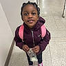 child, girl, smiling, portrait, standing, indoor, hallway, tile_floor, backpack, pink_backpack, braided_hair, jacket, purple_clothing, sneakers, money, dollar_bill, hands, schoolchild, front_facing, happy