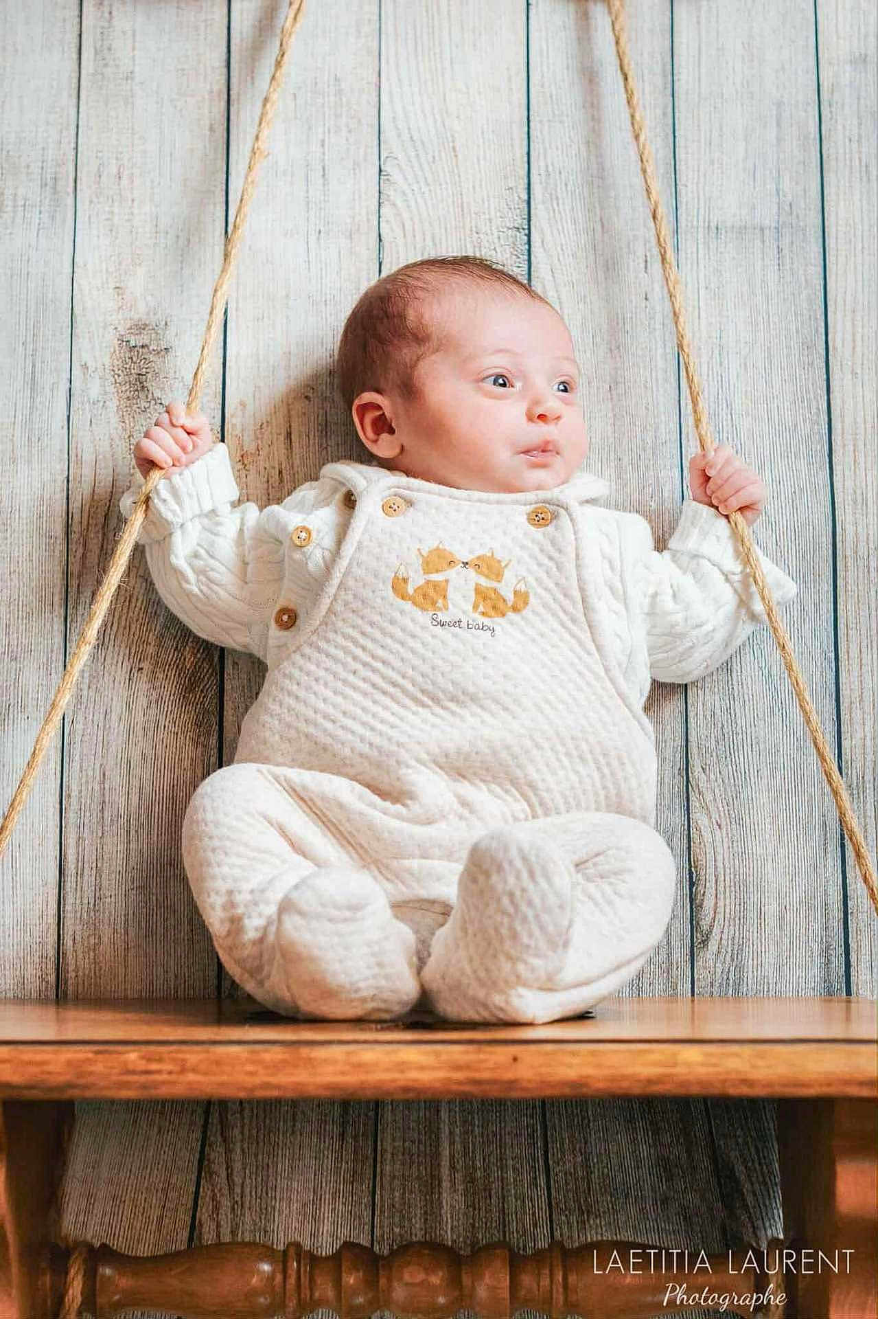 Lola a rejoint le concours — aidez-le/la à gagner de superbes lots ! baby, child, swing, rope, wooden_swing, wooden_background, indoor, white_clothing, cute, infant, seated, portrait, cozy, playful, hands, face, eyes, buttoned_outfit, soft_texture, photography