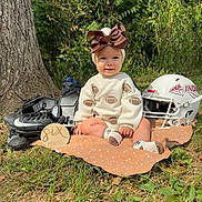 Milana is registered to the contest to win money with this photo: baby, infant, child, six_months, football, football_helmet, football_cleats, bow, headband, sweater, smiling, sitting, portrait, outdoor, woodland, trees, grass, blanket, sports_theme, milestone_photo