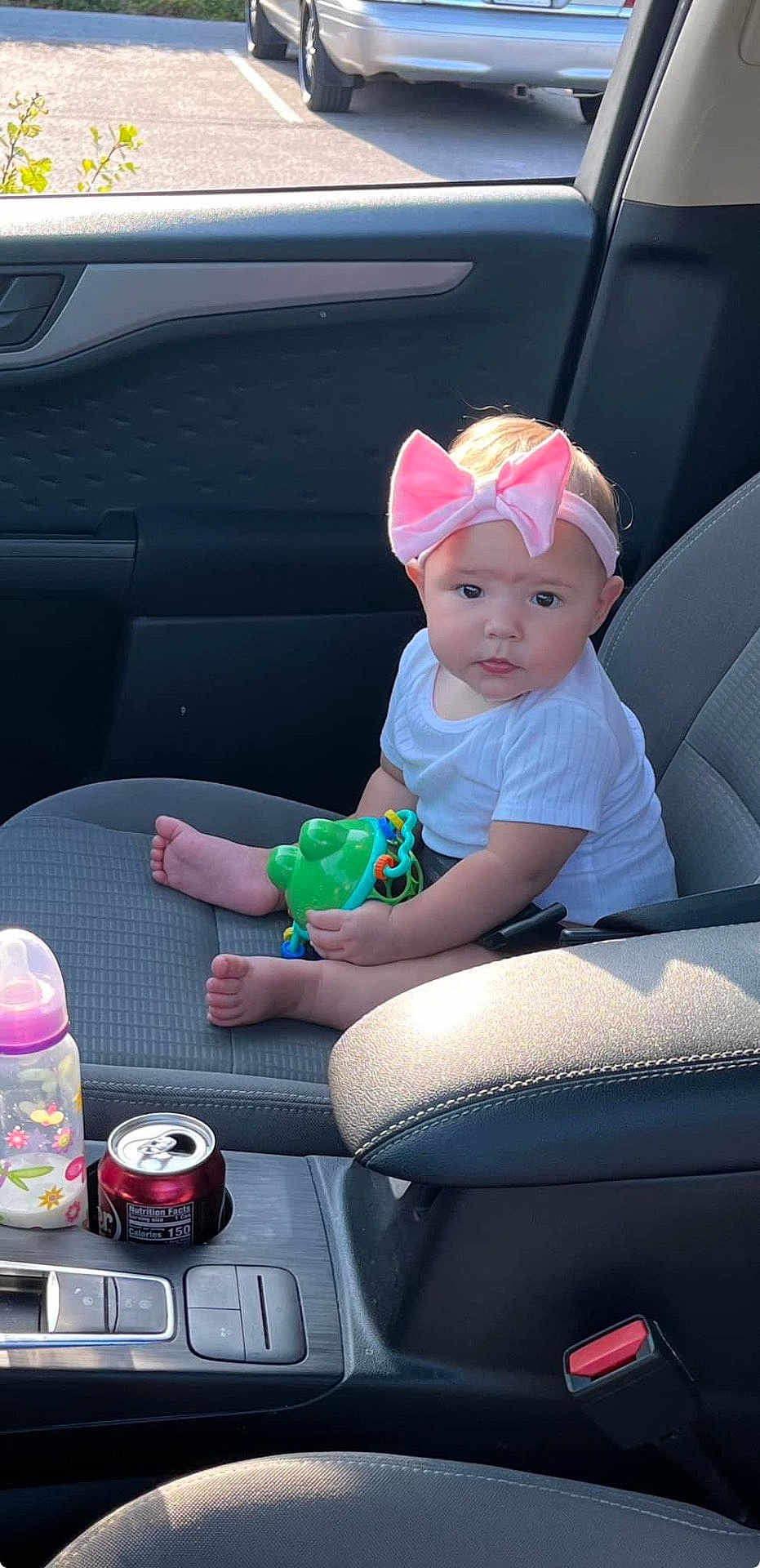 Armani is registered to the contest to win money with this photo: baby, child, car_interior, car_seat, toy, headband, pink_bow, white_shirt, baby_bottle, soda_can, cup_holder, seatbelt, window, parking_lot, sunlight, foot, hand, face, person, infant