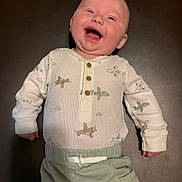Spencer is registered to the contest to win money with this photo: baby, infant, smiling, laughing, onesie, airplane_pattern, buttons, pants, lying_down, mat, closeup, happy, hand, tongue, cheek, bald_head, indoor, pale_skin, clothing, portrait