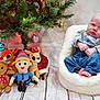 baby, child, plush_toys, christmas_tree, holiday, santa_claus, reindeer, gingerbread_man, stuffed_animal, blue_shirt, suspenders, jeans, white_chair, flooring, decorations, festive, indoor, holiday_lights, expression, portrait