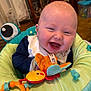 baby, infant, smiling, laughing, toy, bouncer_seat, bib, cute, chubby_cheeks, close_up, indoor, play, happy, portrait, seat, fabric, hand, mouth_open, eyes_squinting, colorful