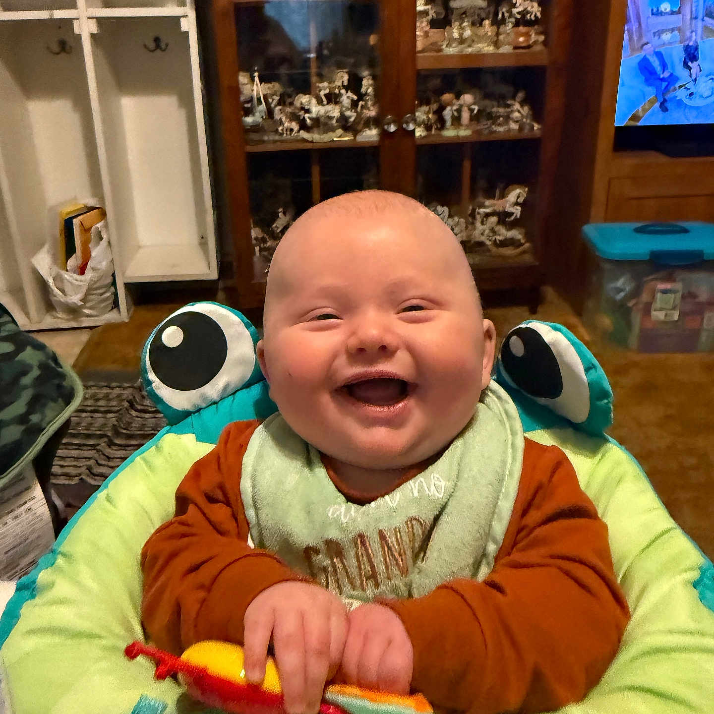 Spencer is registered to the contest to win money with this photo: baby, baby_bouncer, bib, cabinet, cheeks, eyes, furniture, green, hands, happy, indoor, infant, living_room, orange, plush, portrait, seat, smiling, television, toy
