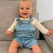 Ethan a rejoint le concours — aidez-le/la à gagner de superbes lots ! baby, child, smiling, denim_overalls, long_sleeve_shirt, couch, fabric, ribbed_texture, indoor, happy, sitting, barefoot, cute, infant, person, portrait, skin, face, hands, feet