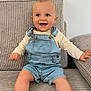 baby, child, smiling, denim_overalls, long_sleeve_shirt, couch, fabric, ribbed_texture, indoor, happy, sitting, barefoot, cute, infant, person, portrait, skin, face, hands, feet