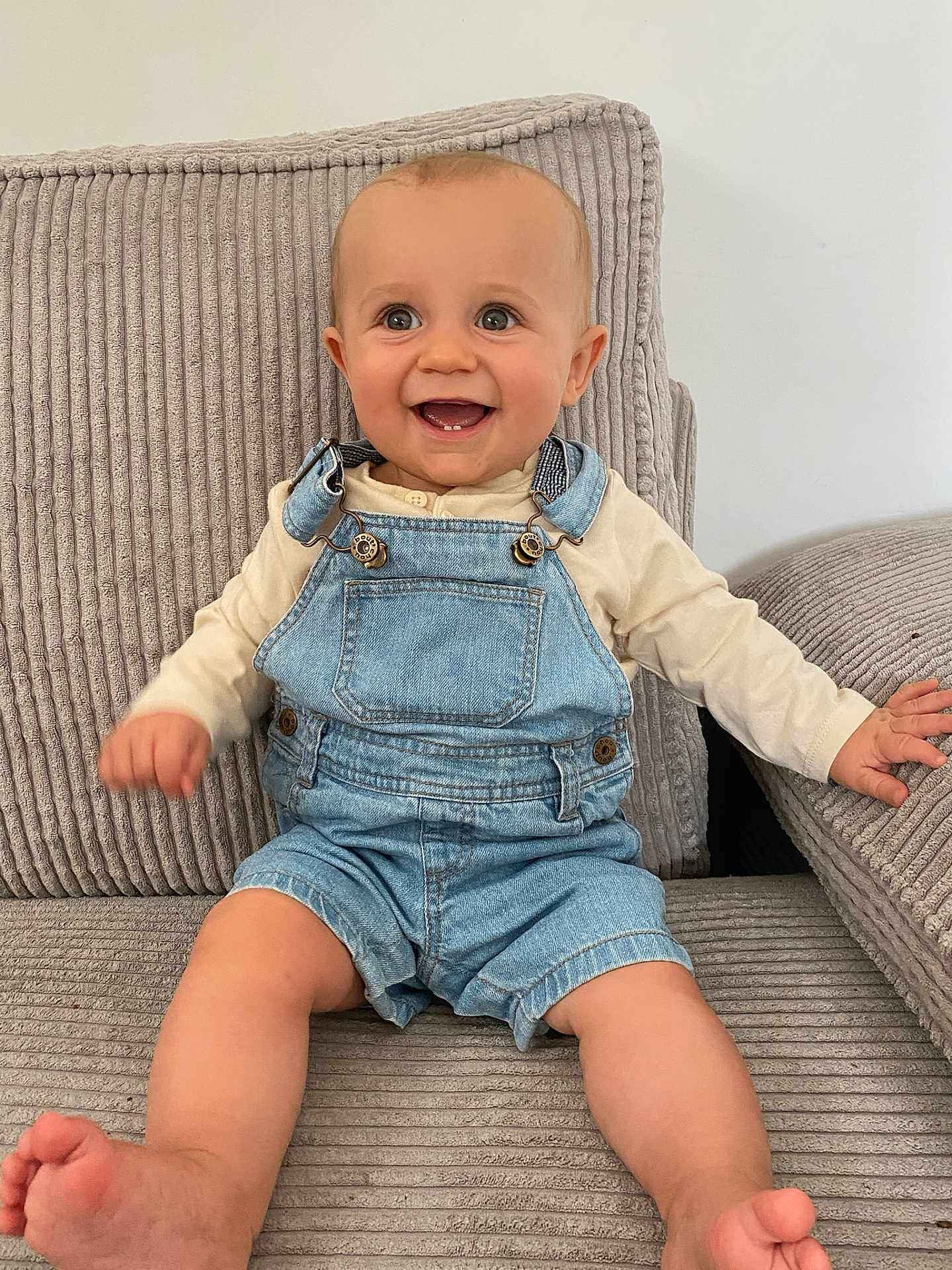 Ethan a rejoint le concours — aidez-le/la à gagner de superbes lots ! baby, child, smiling, denim_overalls, long_sleeve_shirt, couch, fabric, ribbed_texture, indoor, happy, sitting, barefoot, cute, infant, person, portrait, skin, face, hands, feet