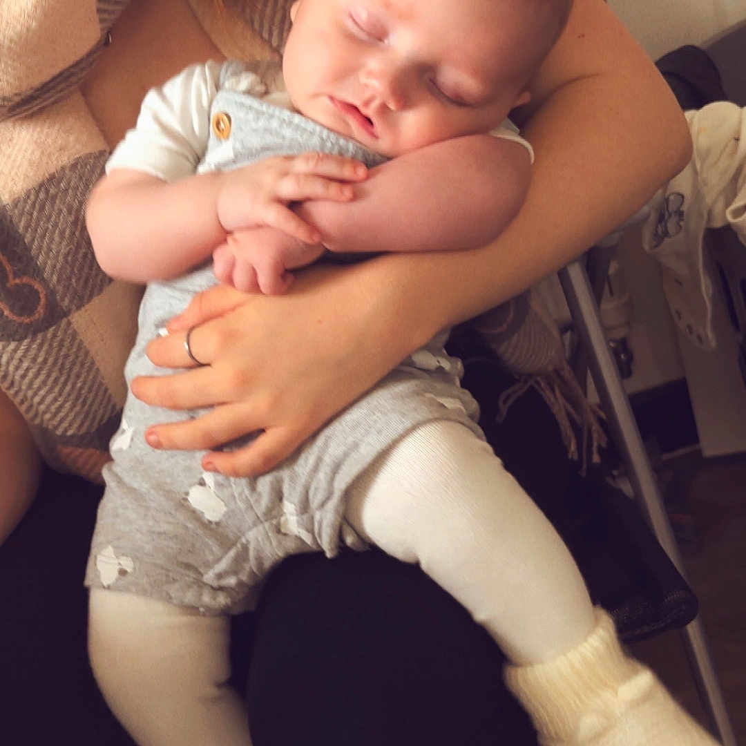 Lyra a rejoint le concours — aidez-le/la à gagner de superbes lots ! adult, arms, baby, blanket, booties, chair, clothing, comfort, cozy, face, hands, holding, home, indoor, peaceful, person, resting, scarf, sleeping, toddler