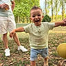 adult, ball, child, daylight, family, grass, hand_holding, happy, leaves, nature, outdoor, person, sandals, shirt, shorts, smiling, summer, toddler, trees, walking