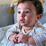 Zayd joined the competition — help win amazing prizes! baby, infant, face, hands, curly_hair, onesie, pillow, teddy_bear_pattern, resting, indoor, closeup, cute, expression, skin, child, portrait, soft_lighting, curious, small_hands, baby_gesture