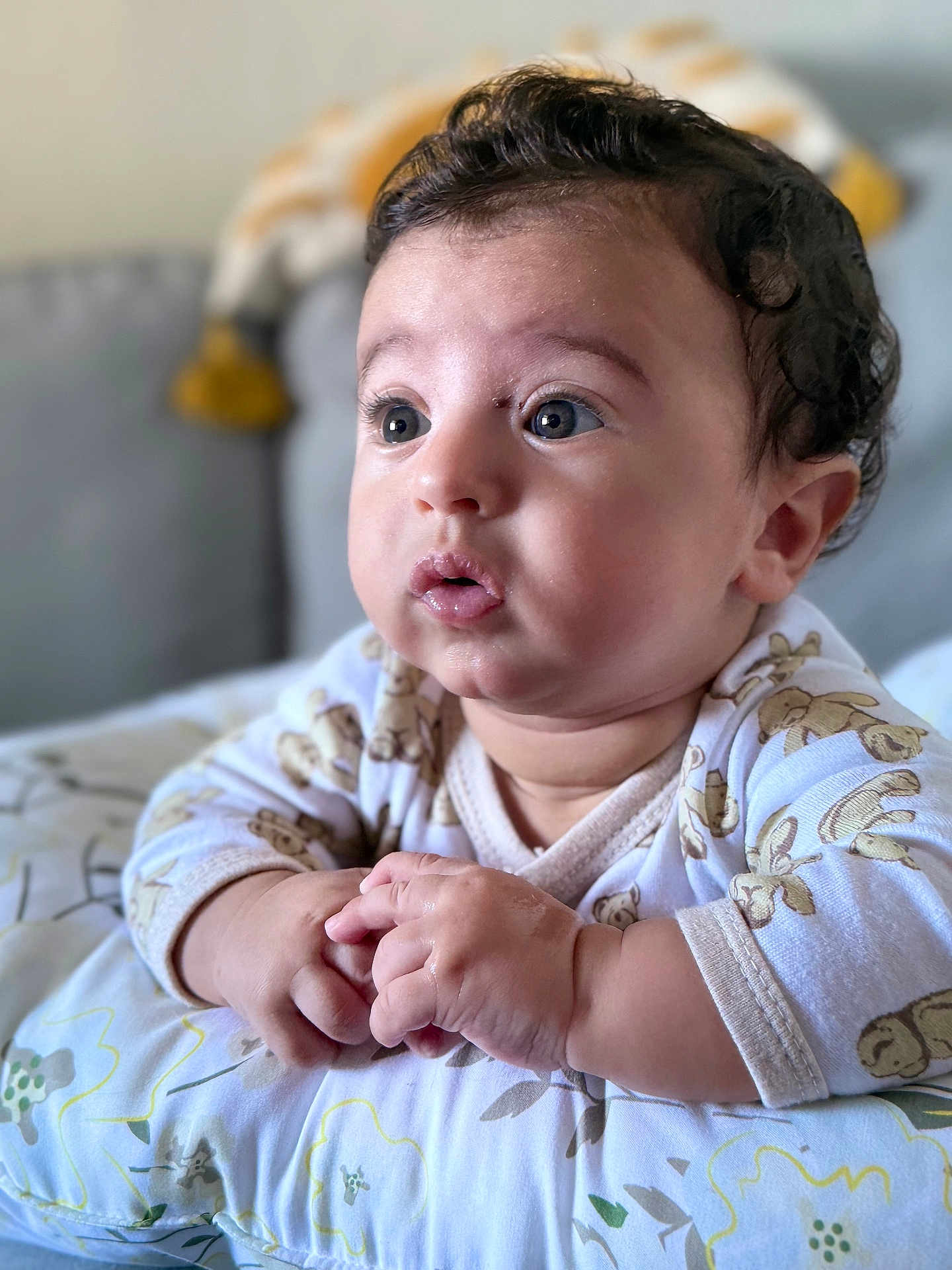 Zayd joined the competition — help win amazing prizes! baby, infant, face, hands, curly_hair, onesie, pillow, teddy_bear_pattern, resting, indoor, closeup, cute, expression, skin, child, portrait, soft_lighting, curious, small_hands, baby_gesture