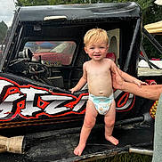 Waylon is registered to the contest to win money with this photo: toddler, child, diaper, race_car, vehicle, outdoor, grass, adult_hand, supporting_hand, black_car, graffiti, red_color, white_color, trees, sky, person, barefoot, smiling, blond_hair, daytime