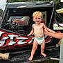 toddler, child, diaper, race_car, vehicle, outdoor, grass, adult_hand, supporting_hand, black_car, graffiti, red_color, white_color, trees, sky, person, barefoot, smiling, blond_hair, daytime