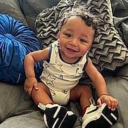 Kdeon is registered to the contest to win money with this photo: baby, child, smiling, curly_hair, onesie, sneakers, couch, pillow, indoor, happy, sitting, footwear, infant, cute, person, portrait, skin, comfort, relaxed, young