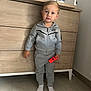 toddler, child, grey_tracksuit, nike, controller, red_controller, barefoot, socks, standing, wooden_dresser, baby_changing_pad, indoor, floor_tiles, curious_expression, short_hair, casual_clothing, person, room_interior, furniture, young_child