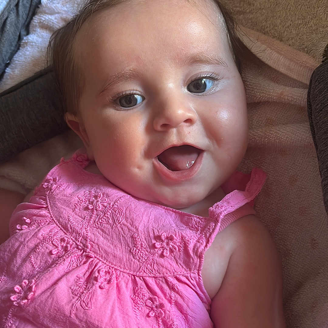 Marie participe au concours pour gagner de l'argent avec cette photo : baby, baby_girl, blanket, child, close_up, clothing, cute, eyes, face, hair_ties, happy, indoors, infant, lying_down, mouth_open, pink_dress, portrait, skin, smiling, soft_texture