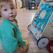 Eloan a rejoint le concours — aidez-le/la à gagner de superbes lots ! toddler, child, indoor, knitted_sweater, green_clothing, wooden_floor, activity_walker, toy, bead_maze, gears, curious_expression, droplet, floor, furniture, blurry_background, baby, candid, sitting, person, playtime