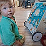 toddler, child, indoor, knitted_sweater, green_clothing, wooden_floor, activity_walker, toy, bead_maze, gears, curious_expression, droplet, floor, furniture, blurry_background, baby, candid, sitting, person, playtime