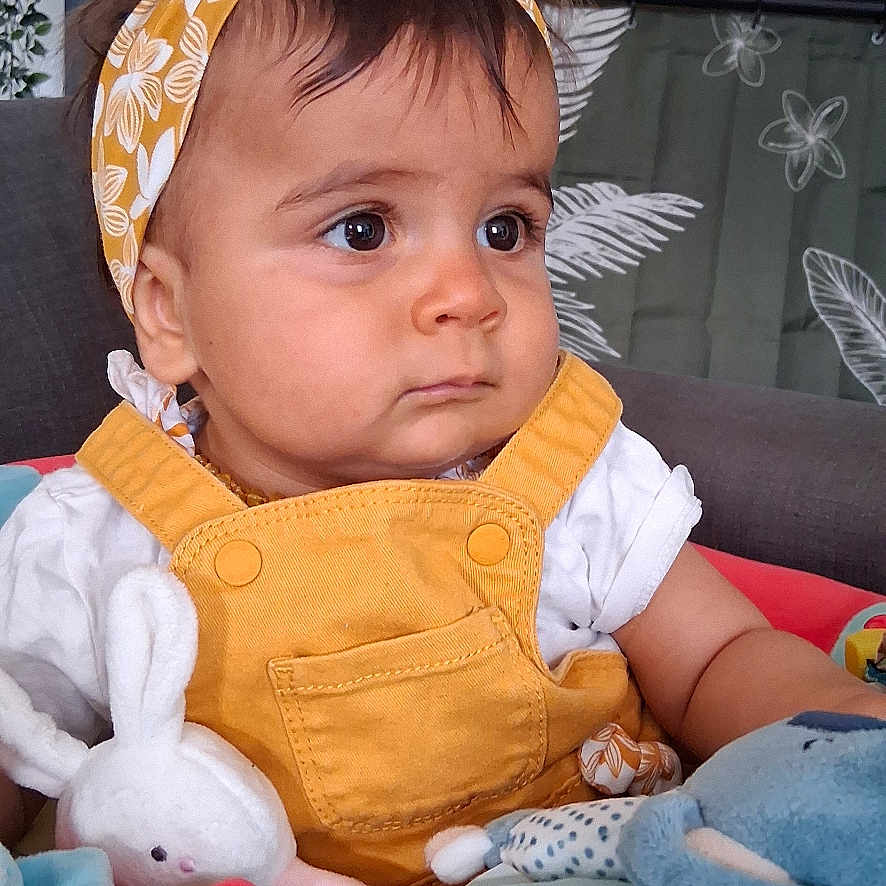 Mia participe au concours pour gagner de l'argent avec cette photo : baby, child, clothing, curious, cute, decor, expression, face, furniture, headband, indoor, overalls, person, plant, plush_toys, portrait, seated, soft_toys, toddler, yellow