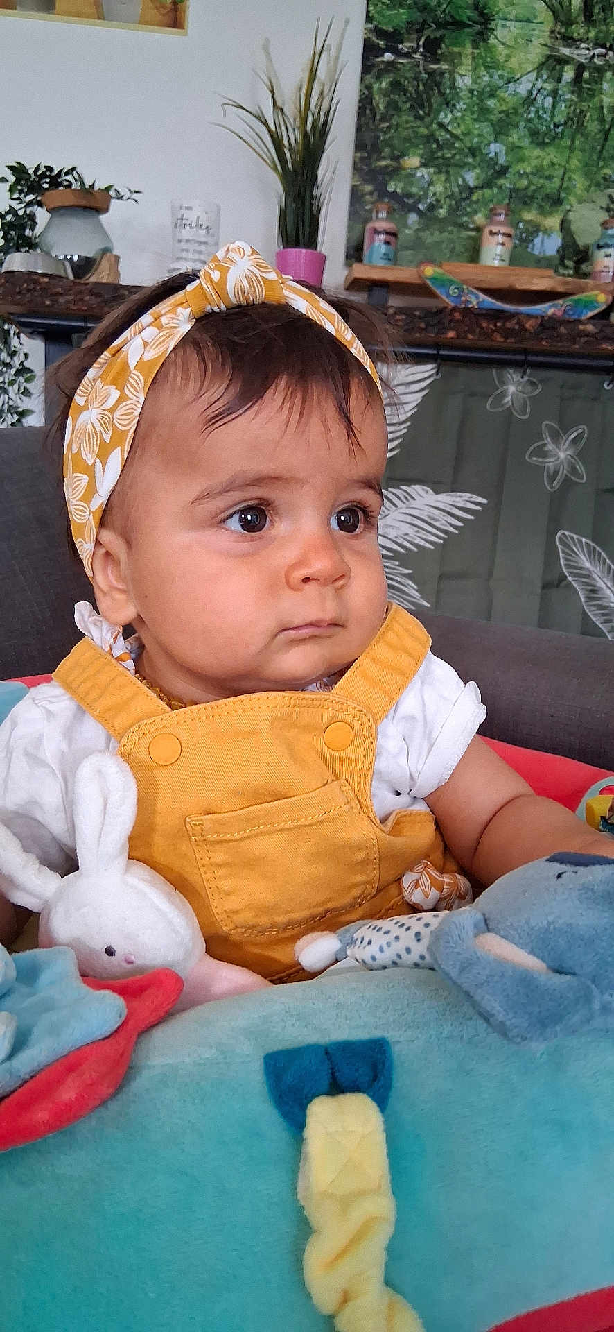 Mia participe au concours pour gagner de l'argent avec cette photo : baby, child, headband, yellow, overalls, plush_toys, indoor, plant, decor, curious, seated, soft_toys, face, clothing, cute, portrait, toddler, furniture, expression, person