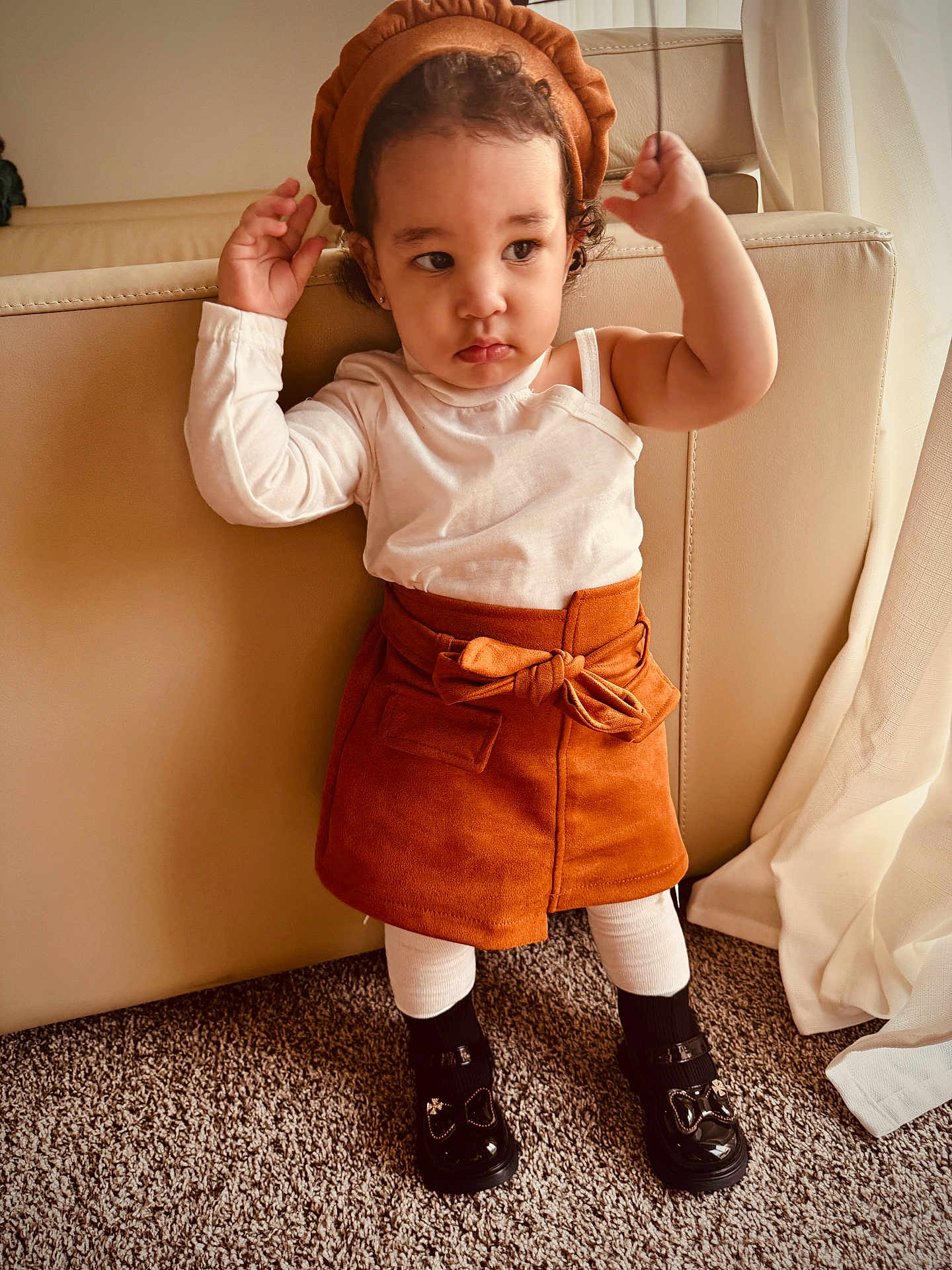 Young Mula is registered to the contest to win money with this photo: toddler, child, standing, beret, skirt, white_top, black_shoes, carpet, couch, indoor, curly_hair, fashion, cute, portrait, person, home, window_curtains, neutral_colors, expression, hands_up