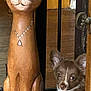 puppy, dog, wooden_statue, cat_statue, floor, wood, necklace, carving, pet, animal, brown, white, cute, indoor, door, door_handle, decor, shiny, eyes, face