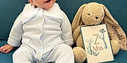 Léopold a rejoint le concours — aidez-le/la à gagner de superbes lots ! baby, infant, onesie, smile, happy, plush_toy, bunny, sign, text, couch, blue_background, sitting, child, cute, portrait, indoor, soft_toy, young_child, buttoned_clothing, collar