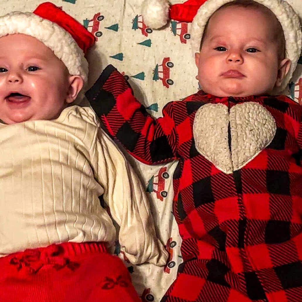 Kenlie N Kylie is registered to the contest to win money with this photo: baby, blanket, christmas_theme, cozy, cream_sweater, cute, face, headwear, holiday_clothing, indoors, infant, lying_down, pattern, plaid, red_and_black, santa_hat, serious_baby, smiling_baby, truck_pattern, two_babies