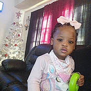 Royalty is registered to the contest to win money with this photo: toddler, child, girl, bow, braids, toy, chair, leather, christmas_tree, ornaments, holiday, indoor, curtain, window, pink, white, black, decor, portrait, casual