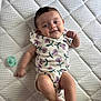baby, infant, smiling, lying_down, onesie, floral_pattern, pacifier, quilted_mattress, child, cute, happy, skin, person, indoors, young_child, resting, baby_clothes, sleeping_area, comfort, playful