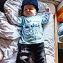 baby, child, hat, blue_shirt, pants, blanket, cushion, bed, indoor, face, person, lying_down, toddler, expression, clothing, resting, toy, casual, headwear, infant