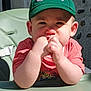baby, toddler, child, green_cap, cap, hands_in_mouth, cute, portrait, highchair, tray, pink_shirt, face, cheeks, ears, eyes, nose, mouth, sunlight, outdoor, stone_wall