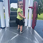 Marceline is registered to the contest to win money with this photo: toddler, child, girl, playground, play_structure, yellow_shirt, black_skirt, sneakers, outdoor, park, trees, metal_floor, steps, smiling, pony_tail, daylight, recreation, happy, person, young_child